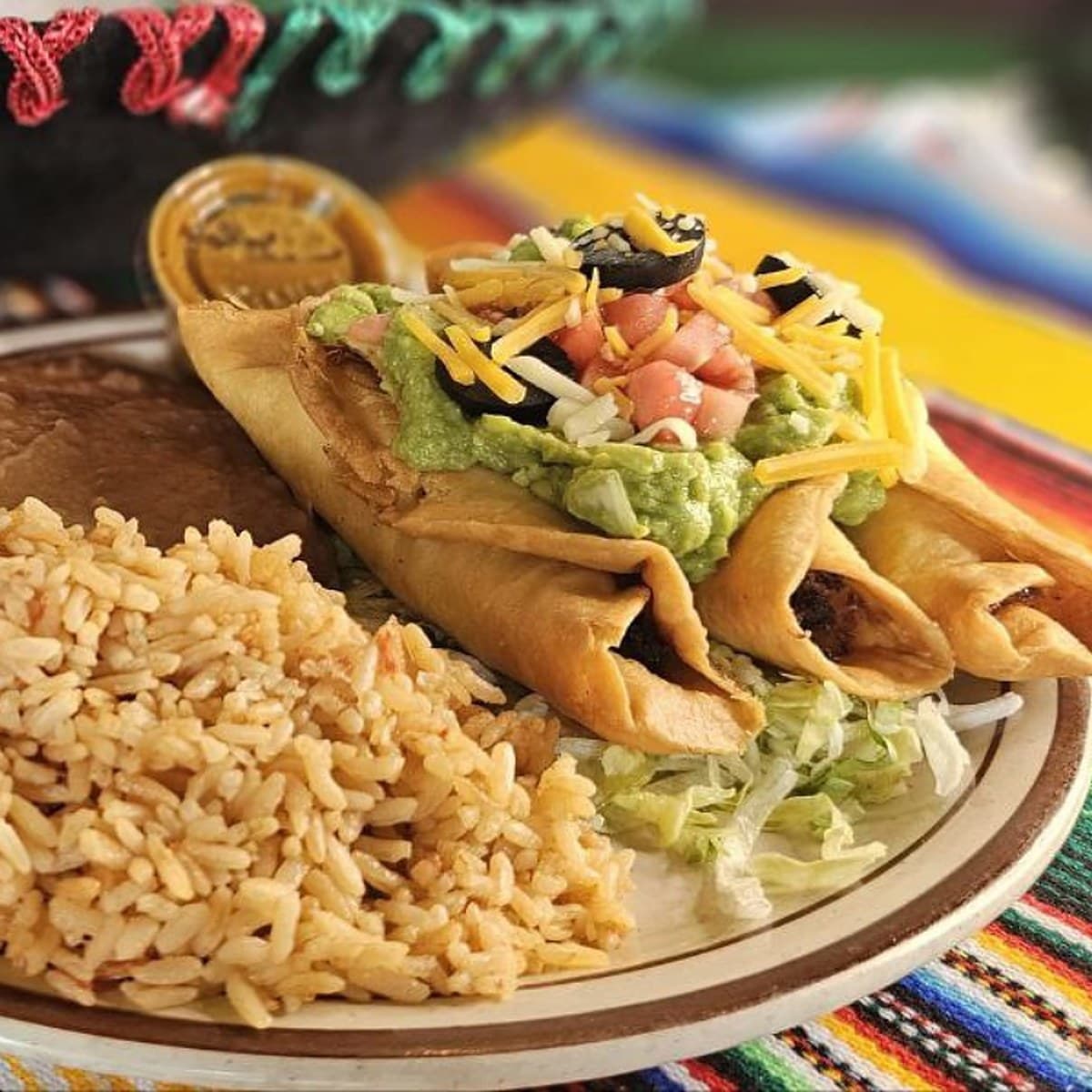 Three crispy flautas topped with guacamole, tomato, cheese and black olives, with Mexican rice, on a serape at Mexican Fiesta 2