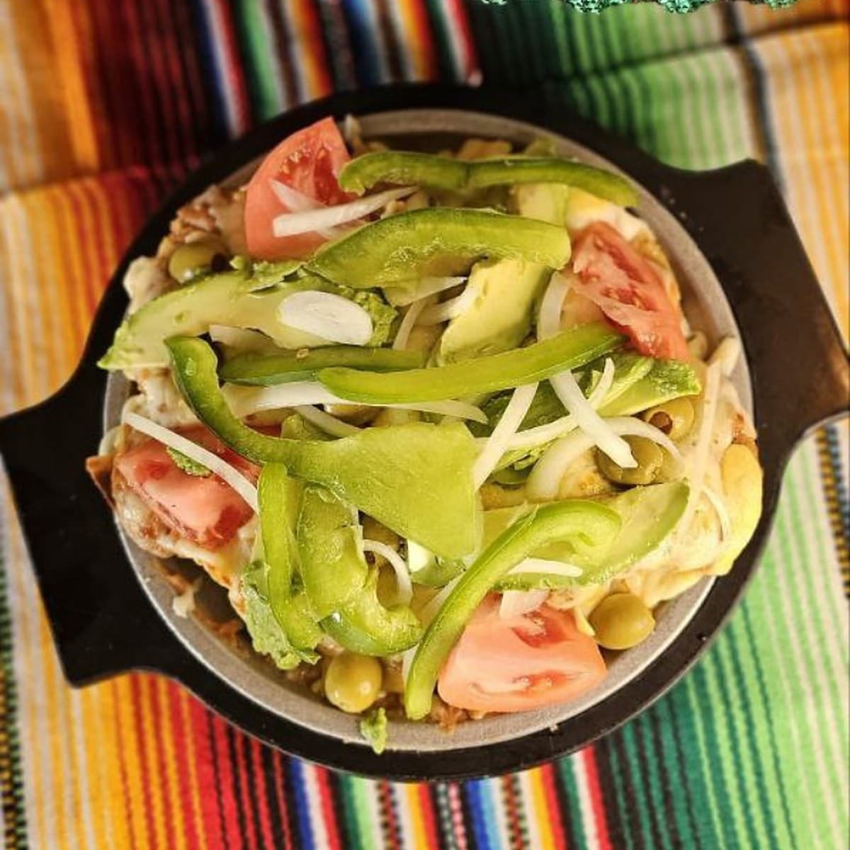 Mini Super Tostada topped with avocado, green pepper, tomato, lettuce and olives in a cast-iron dish